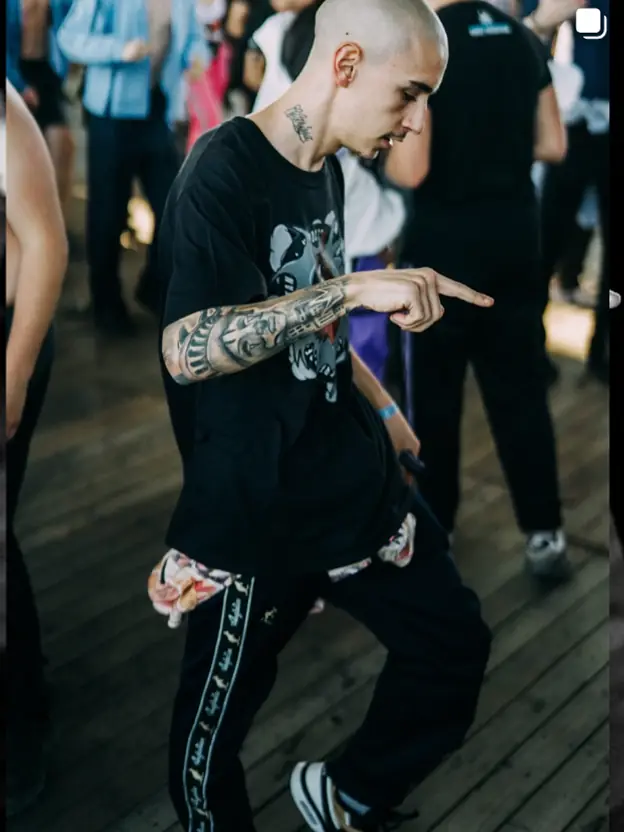 Ninho mid-step on a wooden festival floor, tattooed arm out, daylight party
